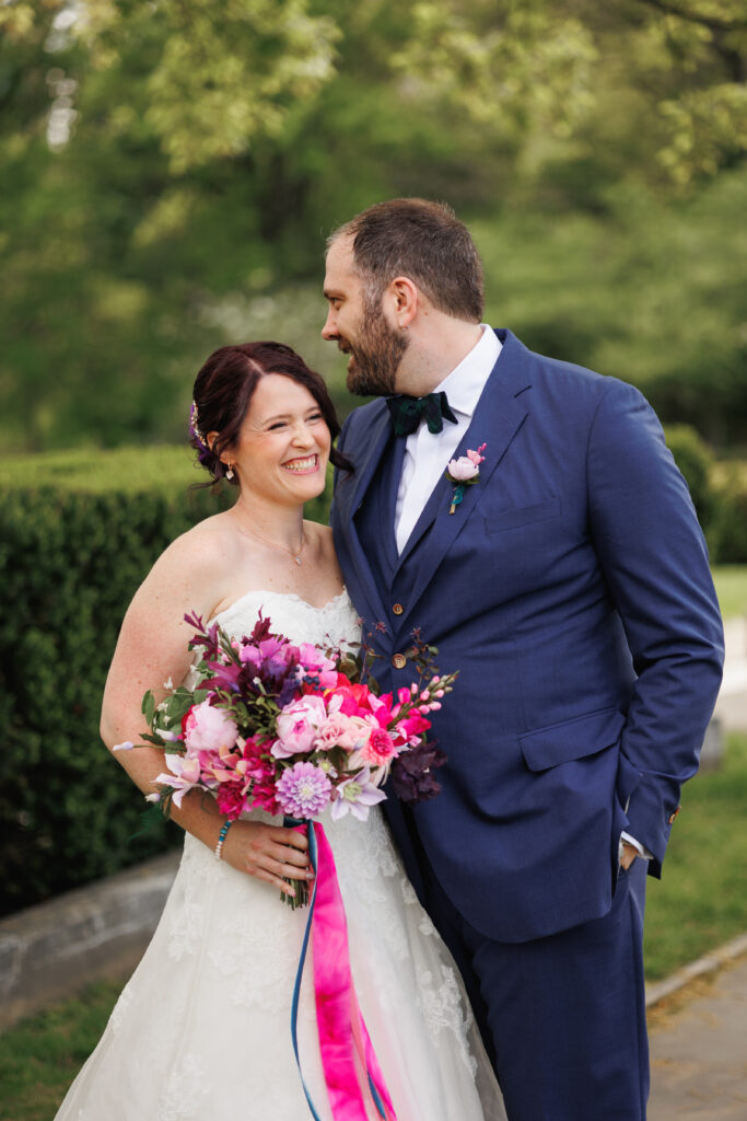 A smiling bride and groom. The bride holds a bouquet of vibrant pink flowers.